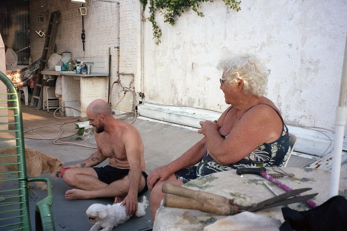 The photo shows two people sitting on chairs outside. One person is holding the leash of a dog, and the other person has their hands on their knees. T