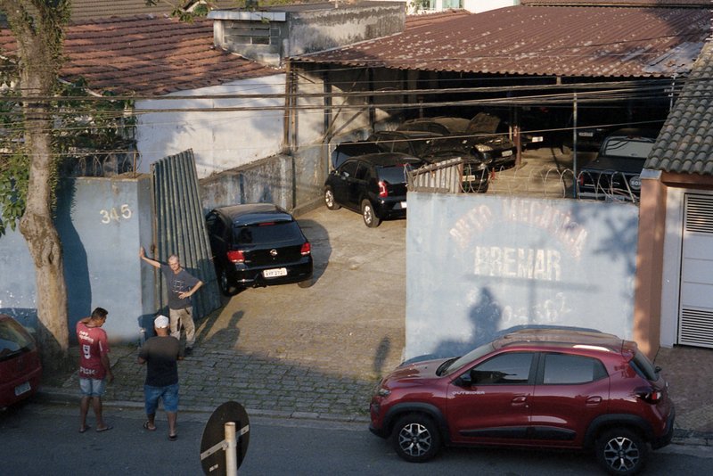 An outdoor scene with several cars parked around a building. There are two individuals standing near the vehicles. The setting appears to be a tropica