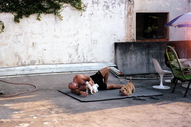 A person is lying on the floor of a dirty room with a dog. Another dog stands nearby, and two more are off-camera. The room has a chair and a hose. Th