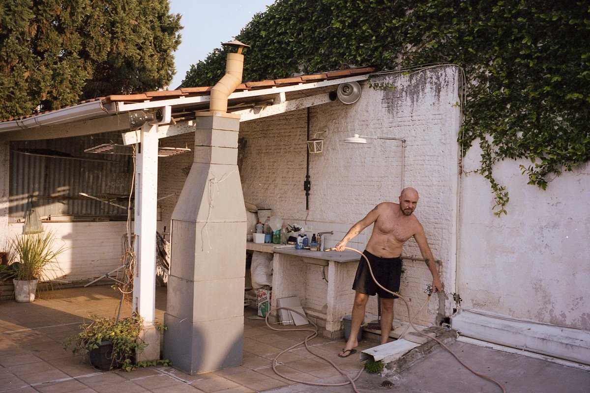 In the photo, a man is engaged in outdoor renovation work. He's standing on a dirt ground near a dilapidated house and appears to be working with elec