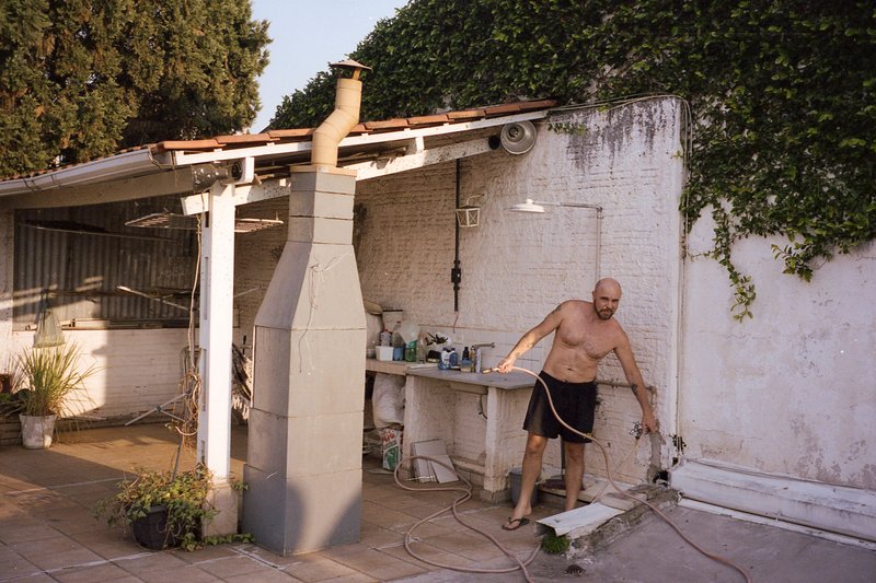 In the photo, a man is engaged in outdoor renovation work. He's standing on a dirt ground near a dilapidated house and appears to be working with elec
