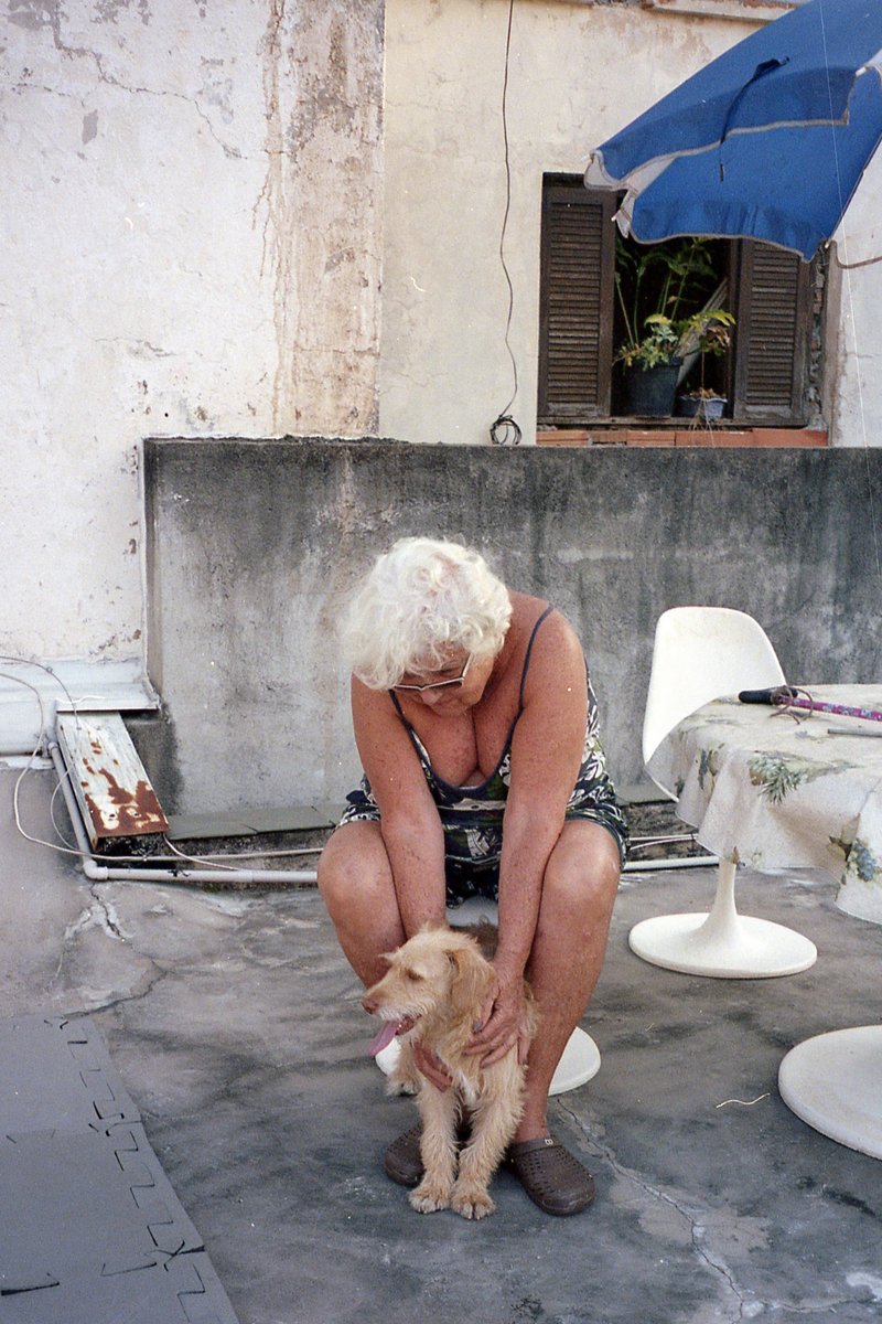 Old woman sitting on ground petting dog. Both are outdoors.
