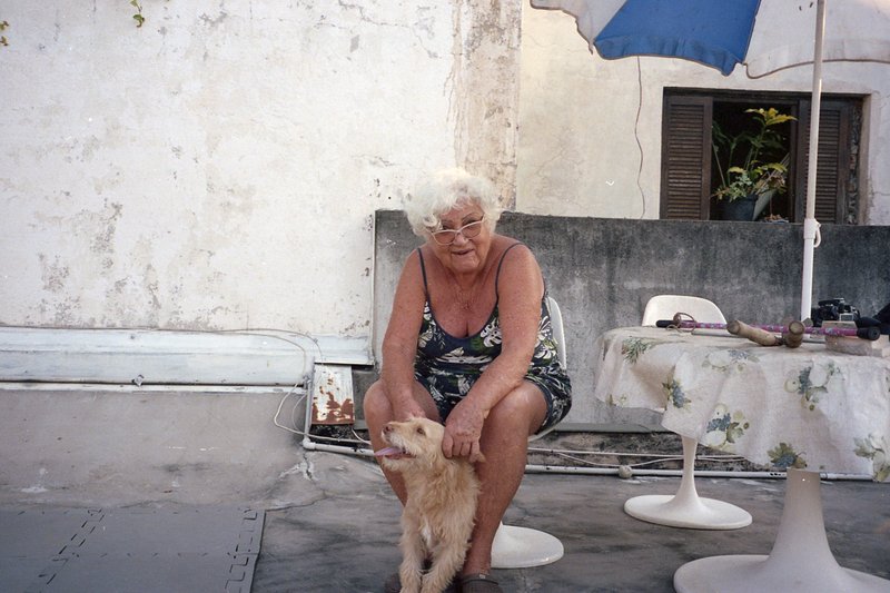 An elderly woman in a summer dress sits on her porch, petting a small dog. She is surrounded by chairs and a dining table, with a blue umbrella provid