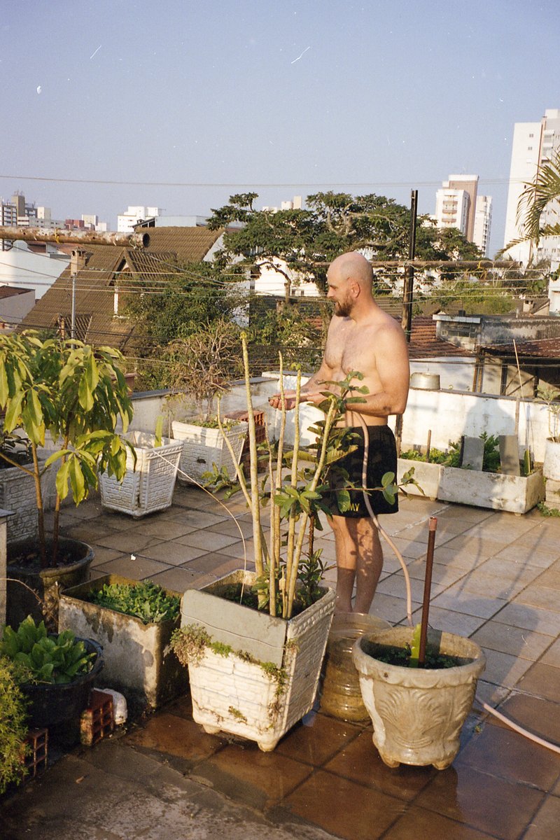 The image is a photograph of a man standing on a tiled floor outdoors. He is shirtless and appears to be looking at some plants. The plants are potted