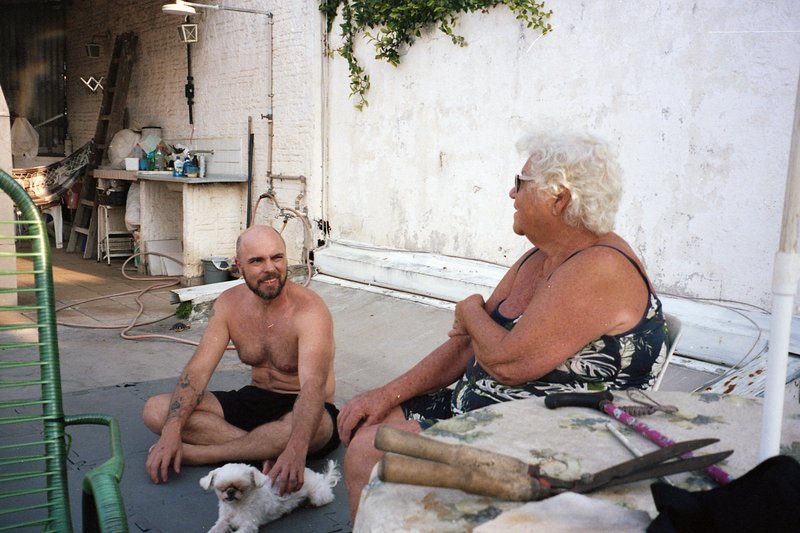 The image shows two individuals, a man and an older woman, sitting on chairs outside. They are engaged in conversation. To their right is a dog standi