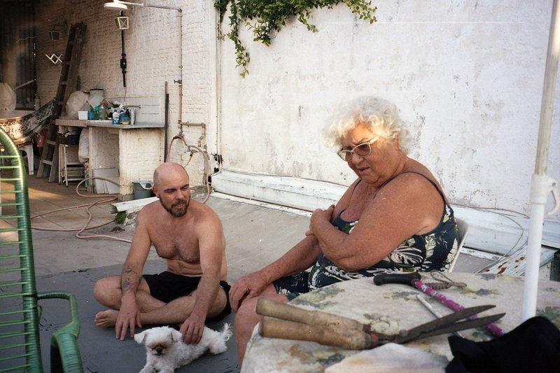Two people sitting on patio chairs. A man kneeling beside them. They are in an outdoor setting with a building and foliage behind them.