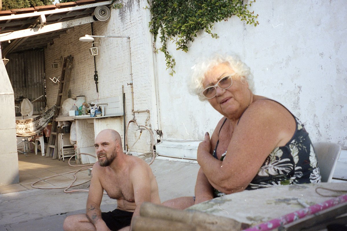 This image is a candid photo of two people sitting on chairs outdoors. They are positioned close to each other, appearing relaxed and engaged in conve