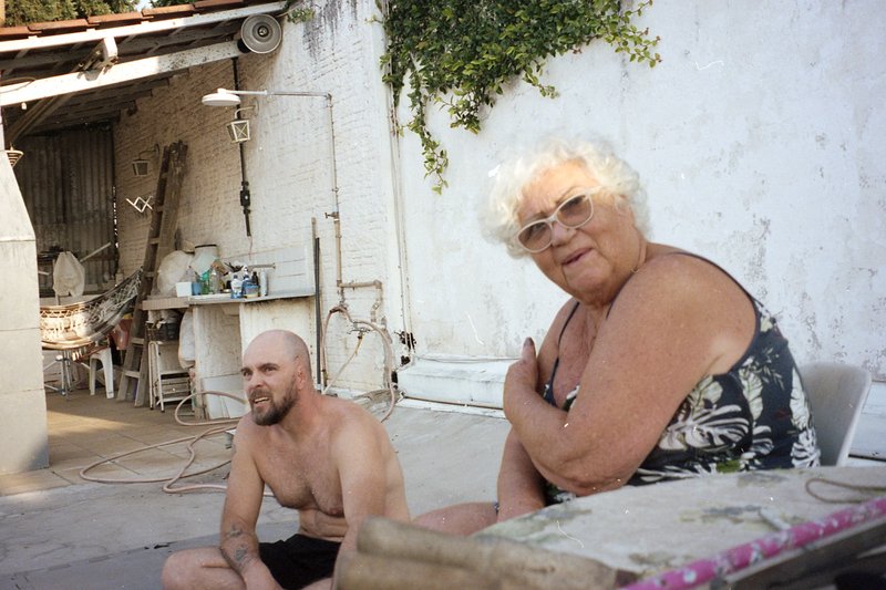 This image is a candid photo of two people sitting on chairs outdoors. They are positioned close to each other, appearing relaxed and engaged in conve