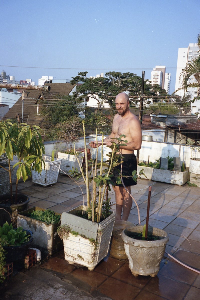 A shirtless man stands in a backyard with potted plants. He holds a water hose and appears to be washing the plants or possibly cleaning himself off.