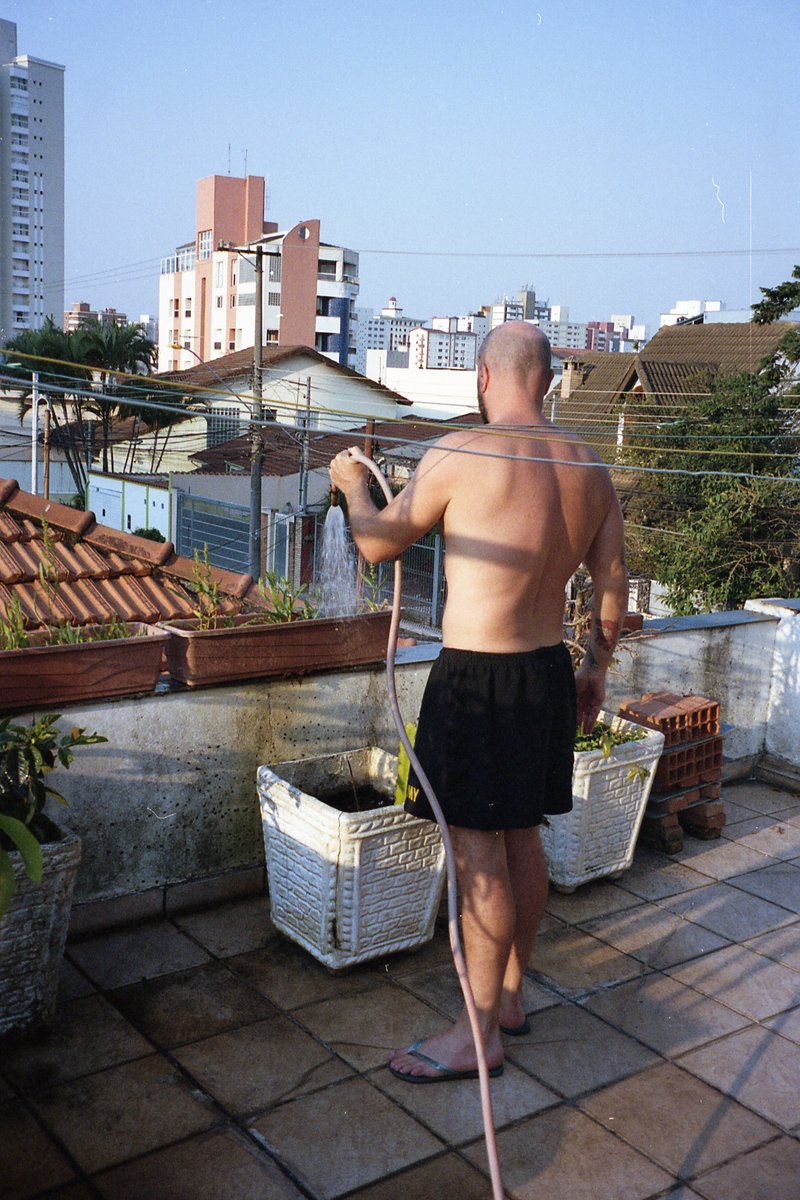 This is a photograph of a man standing on a balcony. He appears to be in the process of washing dishes or clothes, as suggested by the presence of a h