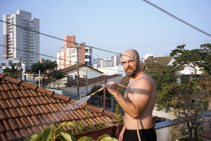 The image shows a man standing on a rooftop. He is shirtless and wearing shorts, indicating it might be warm weather. He is holding a water hose and a