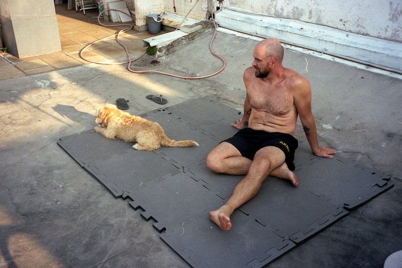 A man is lying on a plastic sheet outdoors. He's shirtless and has his dog next to him. The dog appears to be enjoying the shade beneath them. They se
