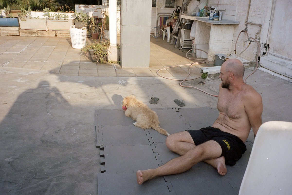 A shirtless man sitting on the ground with his legs stretched out in front of him. He is looking at his dog who is nearby. The man and his dog are bot