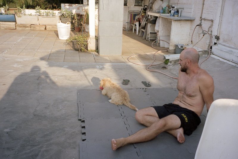 A shirtless man sitting on the ground with his legs stretched out in front of him. He is looking at his dog who is nearby. The man and his dog are bot
