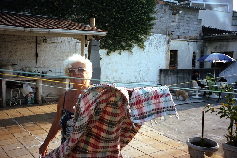This is a color photograph featuring an elderly woman standing on a tiled patio. She has white hair and is wearing a red and black plaid garment. The