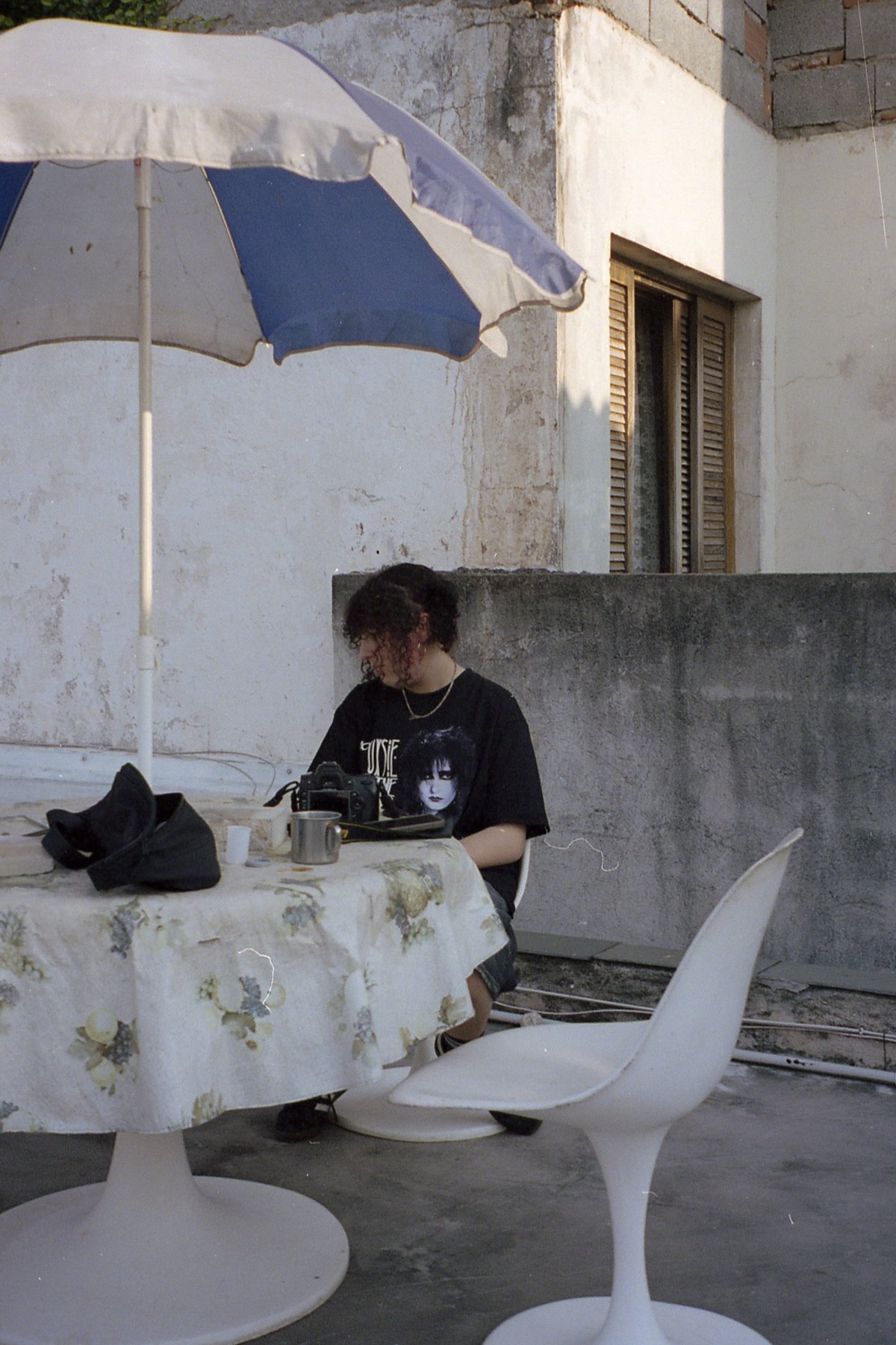 A person sits at a table under a large umbrella on what appears to be a balcony or porch. They seem to be engrossed in something on the table, possibl