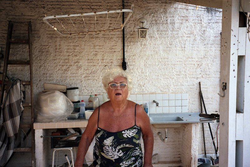 A mature woman stands confidently in front of a makeshift pottery workshop. She's wearing sunglasses and a blue floral top. Behind her, white shelves