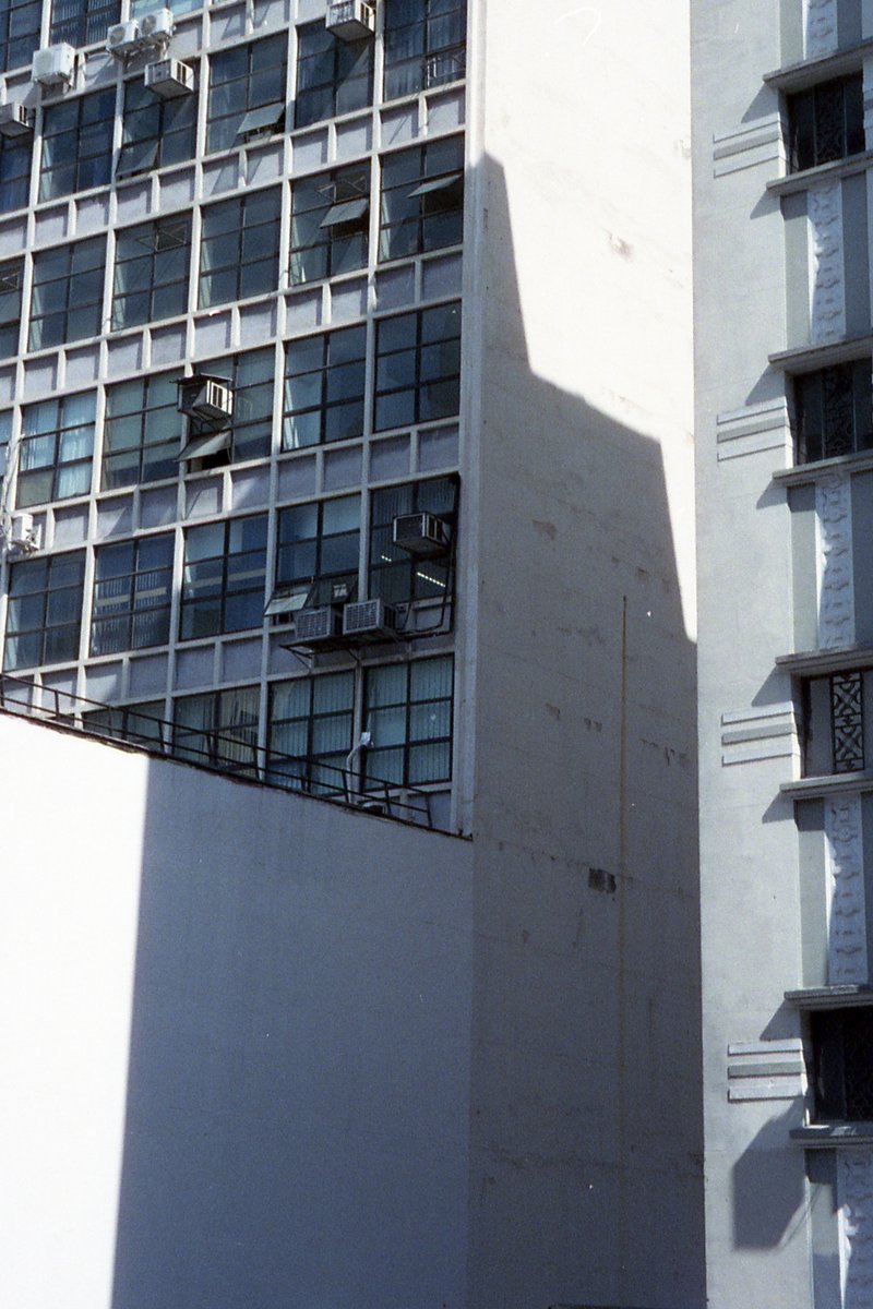 The photo shows a tall residential building. It has a prominent corner balcony on the ground floor with visible air conditioning units. The building's