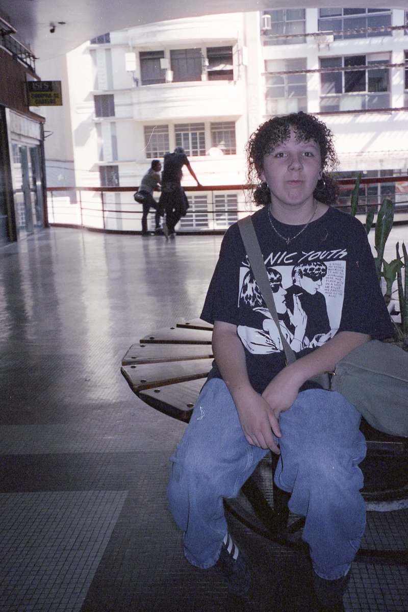 The image depicts a young person seated on a bench indoors. They are wearing a t-shirt with what appears to be text and graphic design. The individual