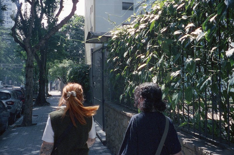 In the image, two individuals are captured in a street scene. The person on the left is donned in a white shirt and has long red hair, while the indiv