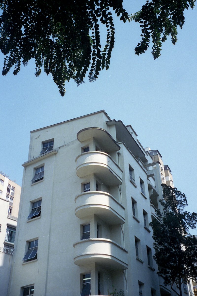 This image features a tall building with balconies, set against a clear sky. The architecture suggests a residential structure, possibly located in a 
