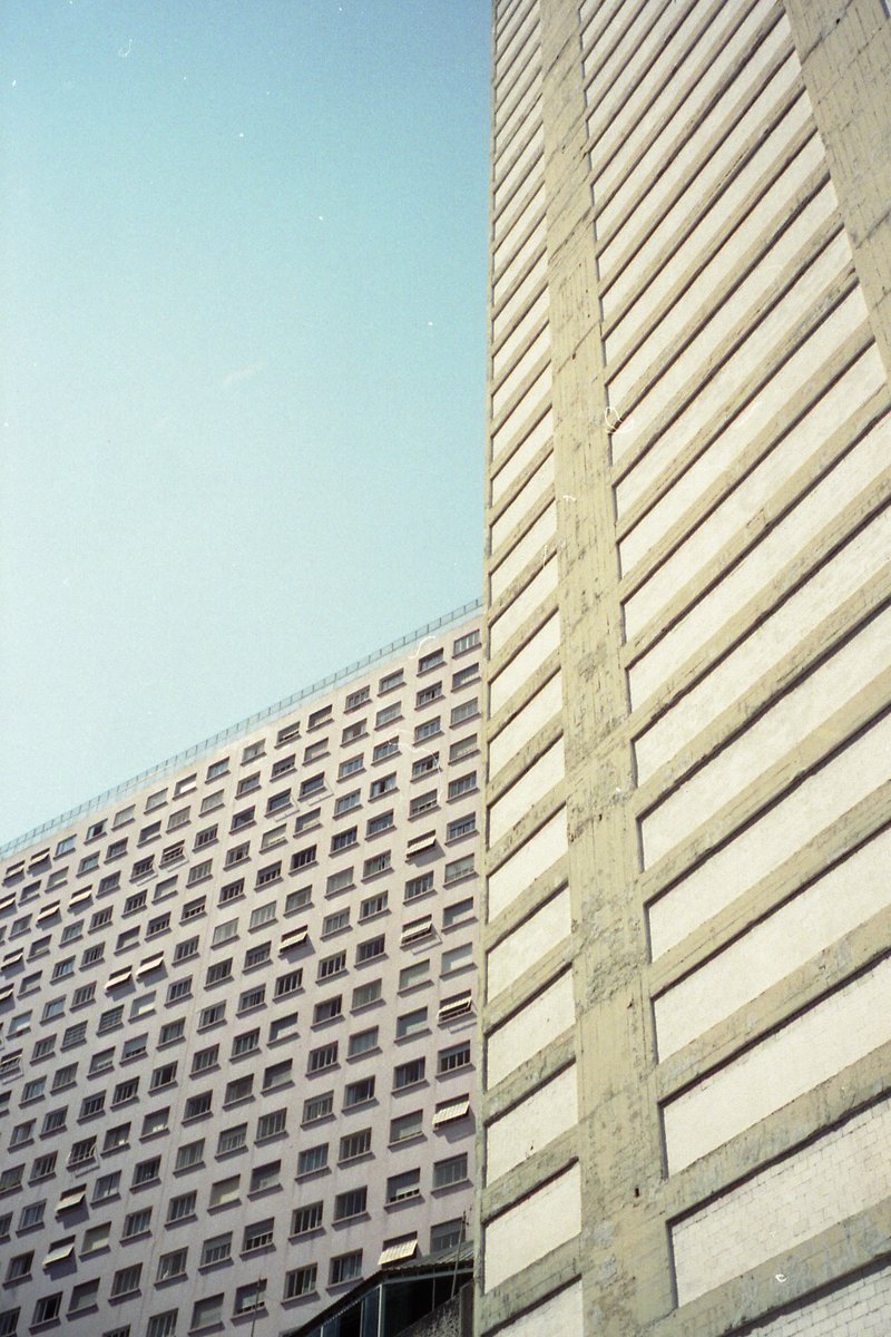 Tall building with multiple windows and a darker base. Another smaller structure adjacent to it. The sky is blue, suggesting daytime.