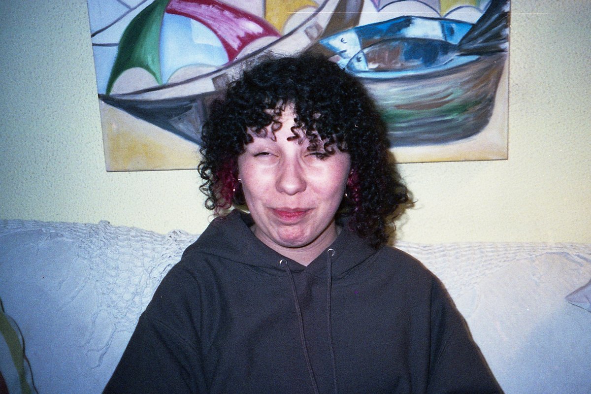 The image is of a young woman with curly hair. She has red eyes and is looking at the camera. Her face appears relaxed, and she is smiling slightly. S