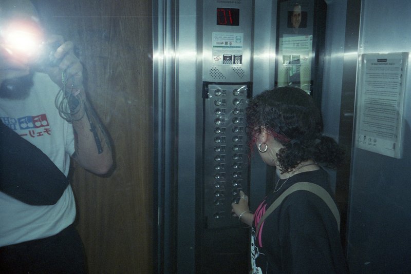 This image depicts a man and woman captured by a photographer in an elevator. They are standing in front of a bank of elevators, with the woman holdin
