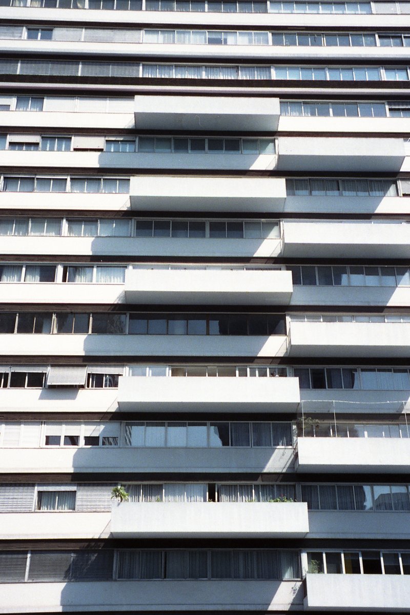 The image depicts a large building with many balconies. It appears to be constructed from concrete or stone blocks, and the balconies have railings al