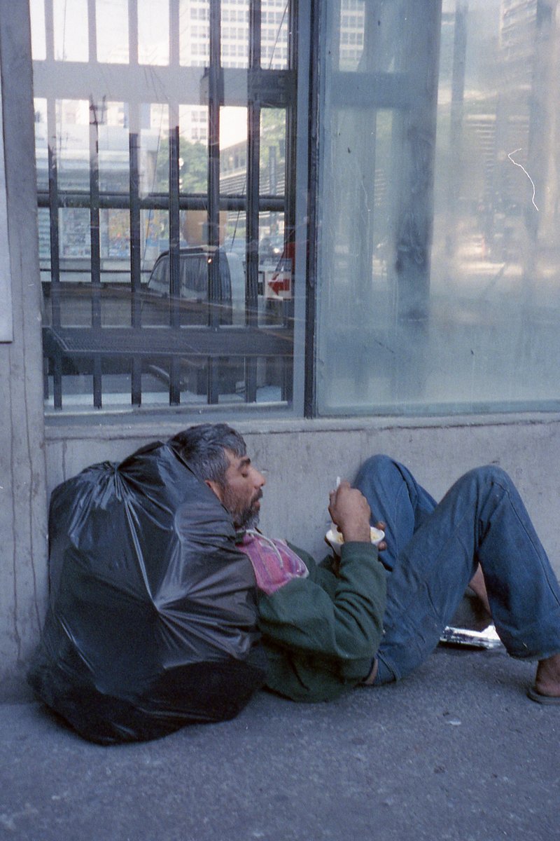 A man is sitting on the ground near a building with bars on the windows. He has a cigarette in his hand and is wearing a black hoodie. The man's belon