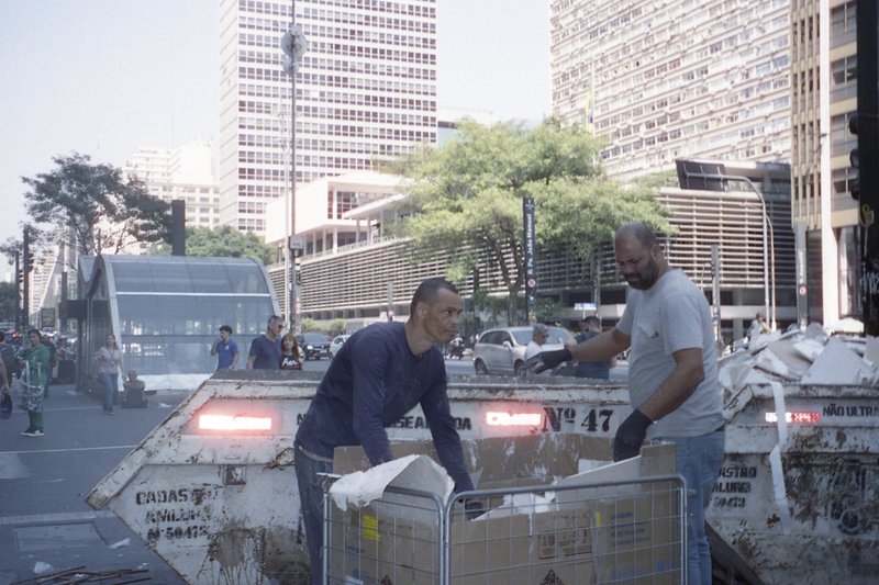 Man loading truck with trash bags. City street scene with buildings and traffic lights in the background. One man is near the truck while another stan