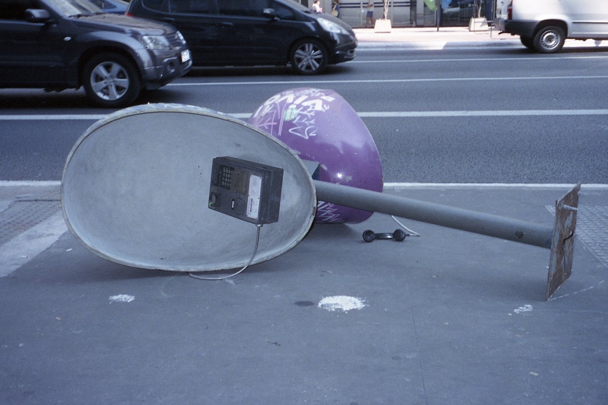 The image features a whimsical scene on a sidewalk. There is an oversized satellite dish lying on its side next to a purple ball. A small car is also 