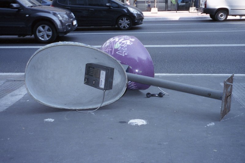 The image features a whimsical scene on a sidewalk. There is an oversized satellite dish lying on its side next to a purple ball. A small car is also 