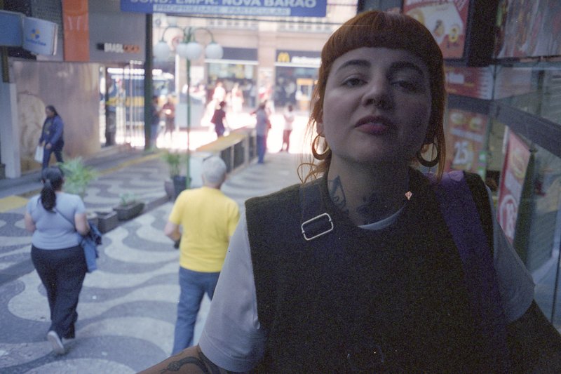 Woman standing in a shopping mall. She has red hair and a nose ring. She is looking away from the camera. The background shows a bustling street scene