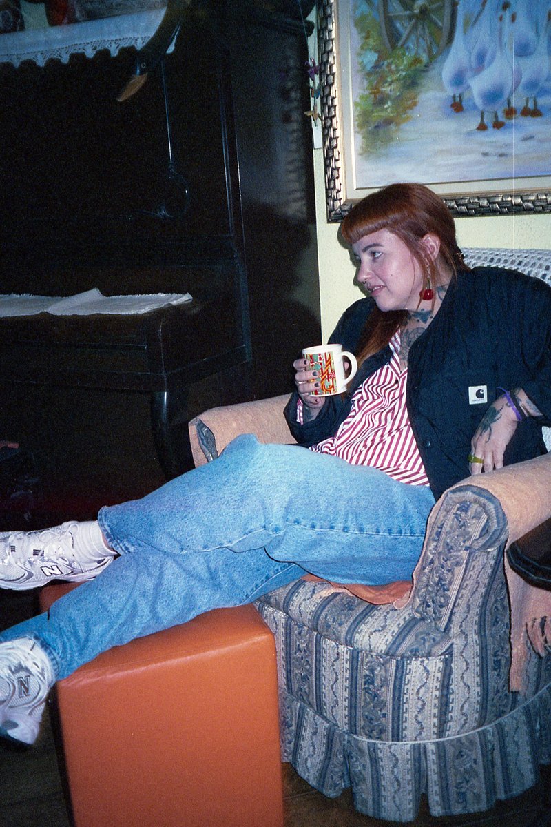 A person with red hair sits on a chair in a room. They are wearing a black jacket over a striped shirt and blue jeans. The individual holds a coffee m