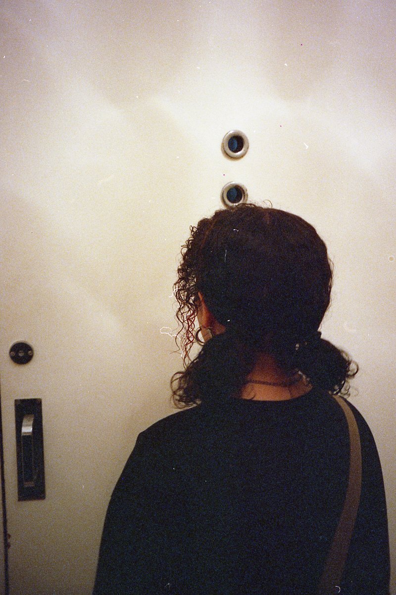 The image is a black and white photograph of a person standing near an elevator. The individual appears to be waiting or preparing to enter the elevat