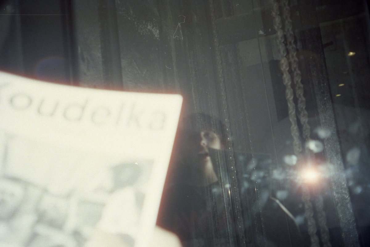 The image shows an out-of-focus photograph of someone holding a book with the word "Pudaka" on it. In the background, there's a window with raindrops 