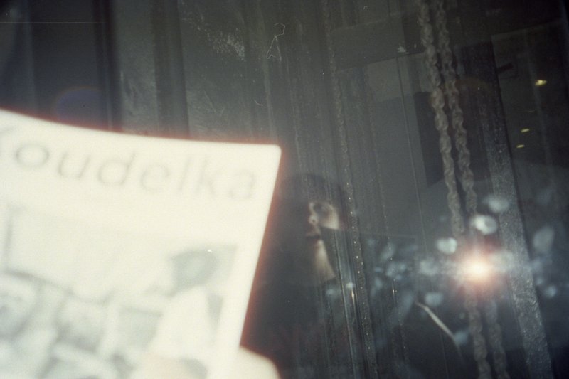 The image shows an out-of-focus photograph of someone holding a book with the word "Pudaka" on it. In the background, there's a window with raindrops 