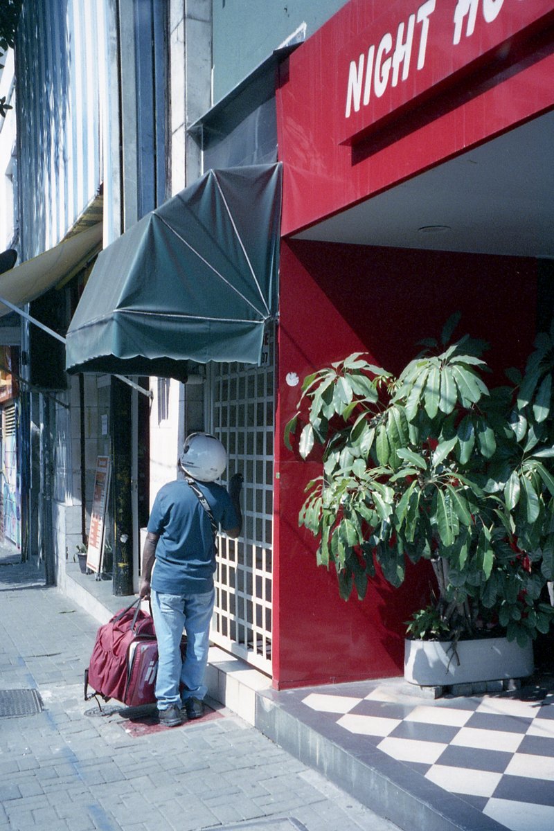 The image features a man standing on the sidewalk next to a red building with "NIGHT HOUSE" written in white. He is wearing a black shirt and has a ba