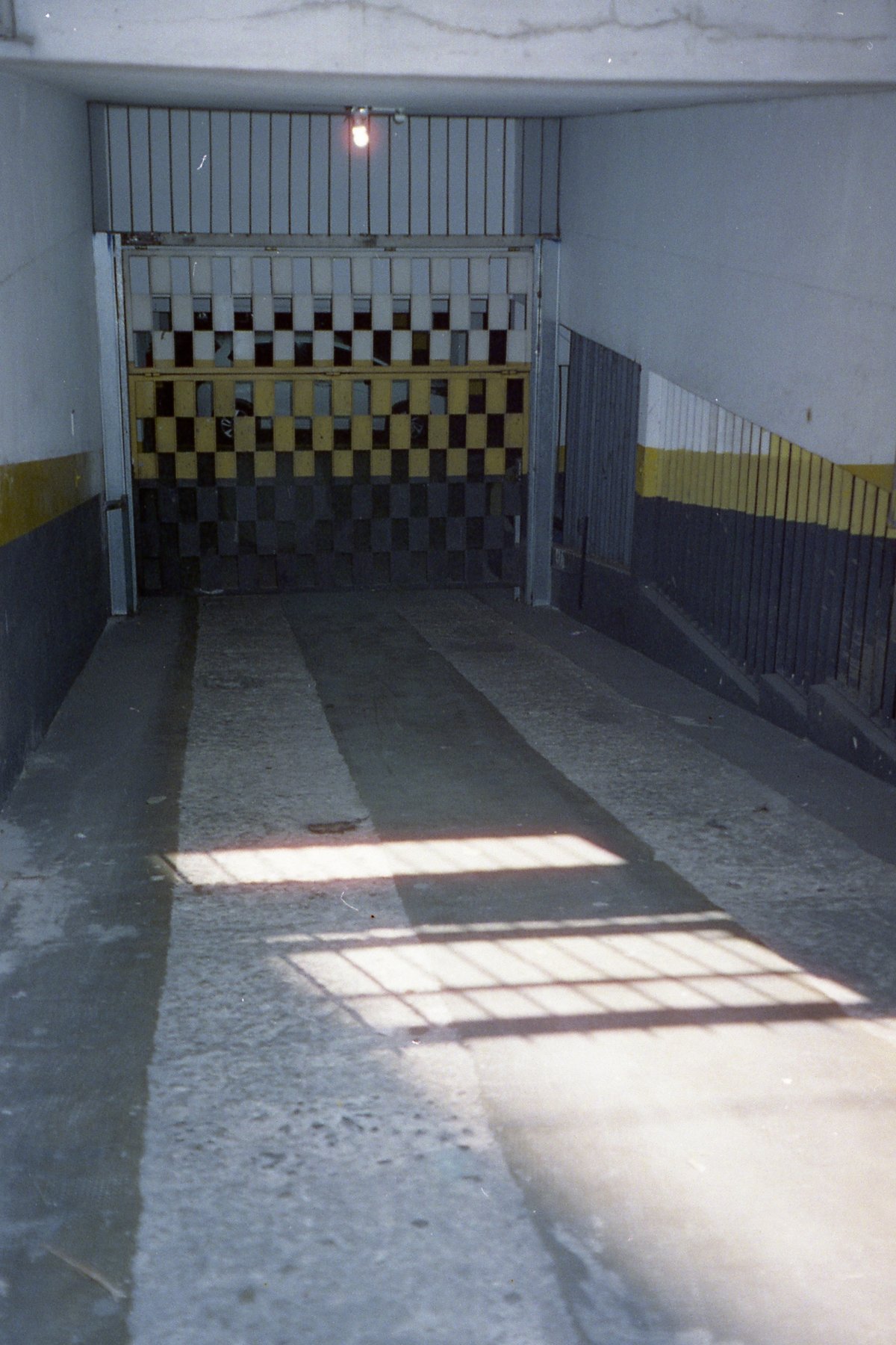 The image depicts the interior of a small room with a yellow and black checkered floor. On one side, there's a gray metal gate or wall. The room appea