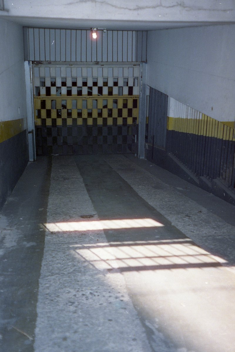 The image depicts the interior of a small room with a yellow and black checkered floor. On one side, there's a gray metal gate or wall. The room appea