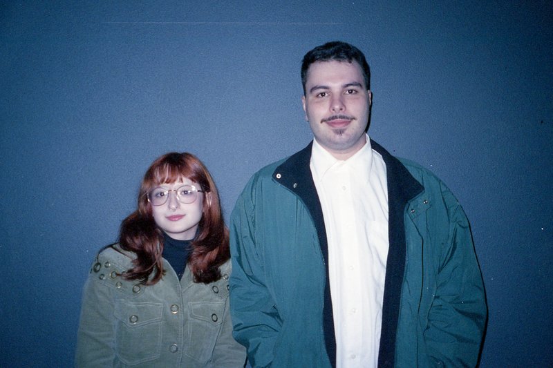The image shows a man and a woman standing closely together. They are both dressed casually, with the man wearing a green jacket and the woman in a wh