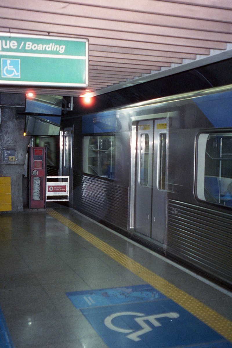 Empty subway station at night with trains on tracks, blue sign, escalator, ticket booth, and a person in the distance.