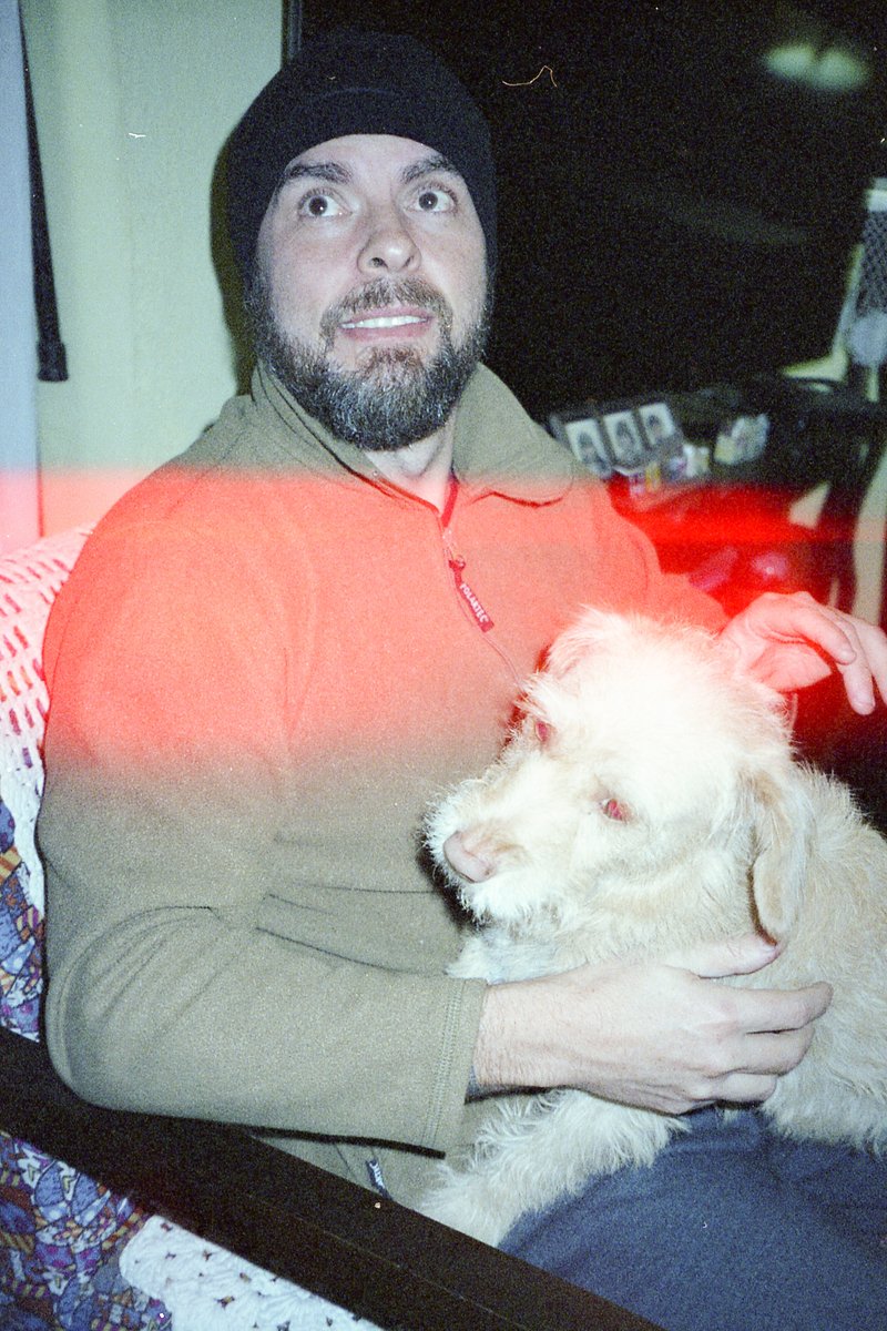 This is a photo of a man sitting and holding a dog. The man appears to be middle-aged with short hair and facial stubble. He's wearing a light brown j