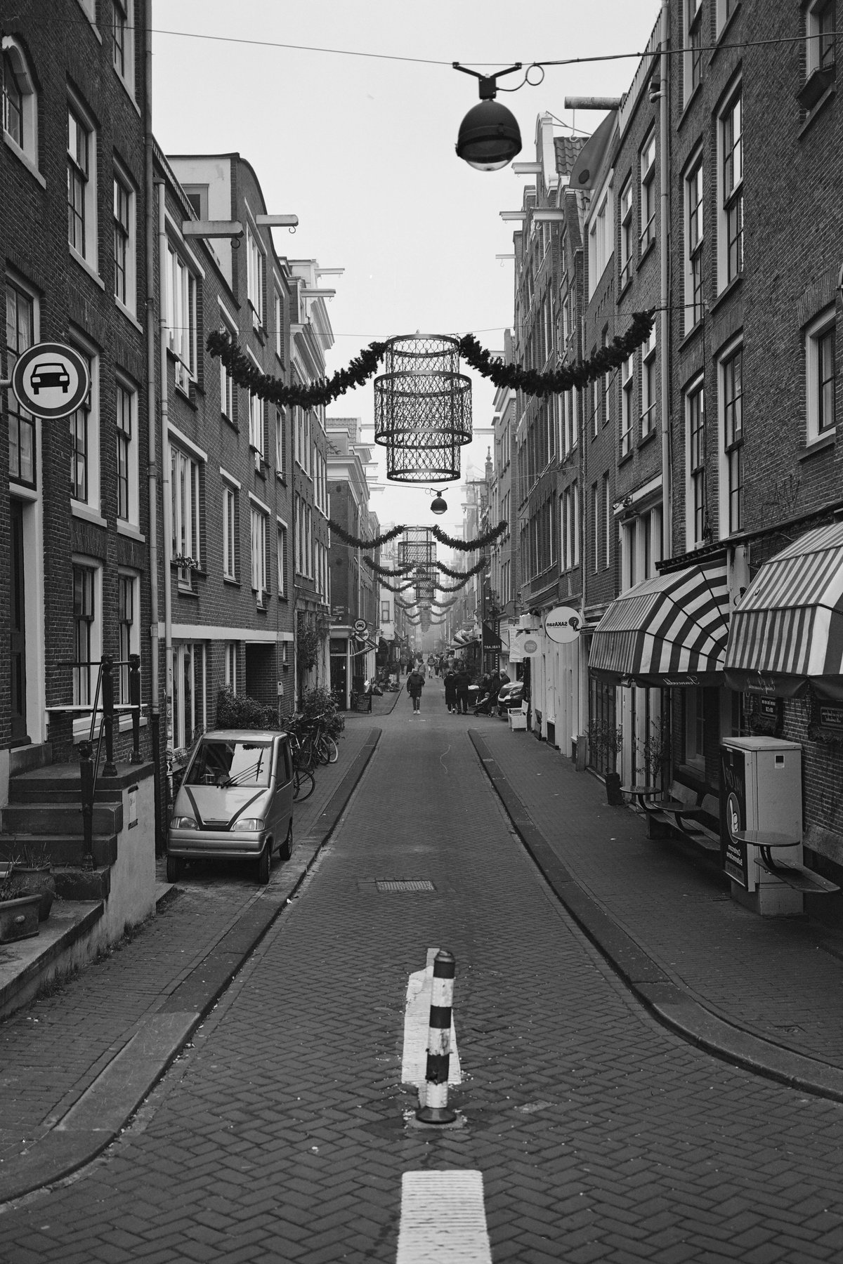  This is a black and white photograph depicting an old European street lined with buildings that have large windows and small doors. The street is narrow and paved with cobblestones, and there are many lights strung up between the buildings, possibly indicating a festive or celebratory occasion. On the right side of the image, there's a sidewalk with some signage, while parked cars can be seen on the left. There is also an elevated train track running parallel to the street. The photo captures the essence of a quaint European town or city alley, giving a sense of being in a historical setting. 