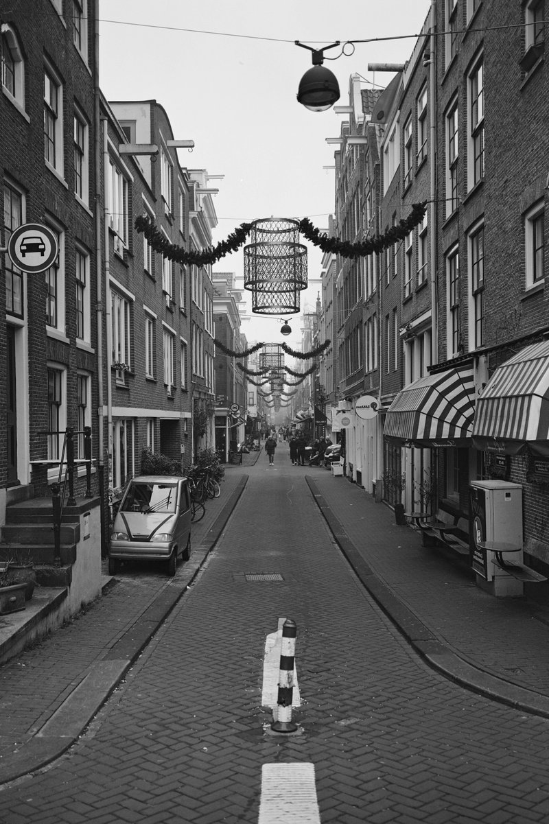 This is a black and white photograph depicting an old European street lined with buildings that have large windows and small doors. The street is narrow and paved with cobblestones, and there are many lights strung up between the buildings, possibly indicating a festive or celebratory occasion. On the right side of the image, there's a sidewalk with some signage, while parked cars can be seen on the left. There is also an elevated train track running parallel to the street. The photo captures the essence of a quaint European town or city alley, giving a sense of being in a historical setting.