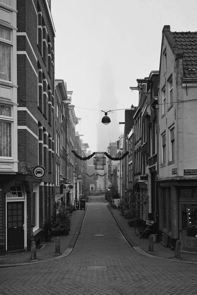 The image is a black and white photograph showcasing a city street lined with tall buildings of various architectural styles. Hanging lights are visible, adding to the urban atmosphere. A solitary figure can be seen walking on the left side of the street, creating a sense of scale and activity.