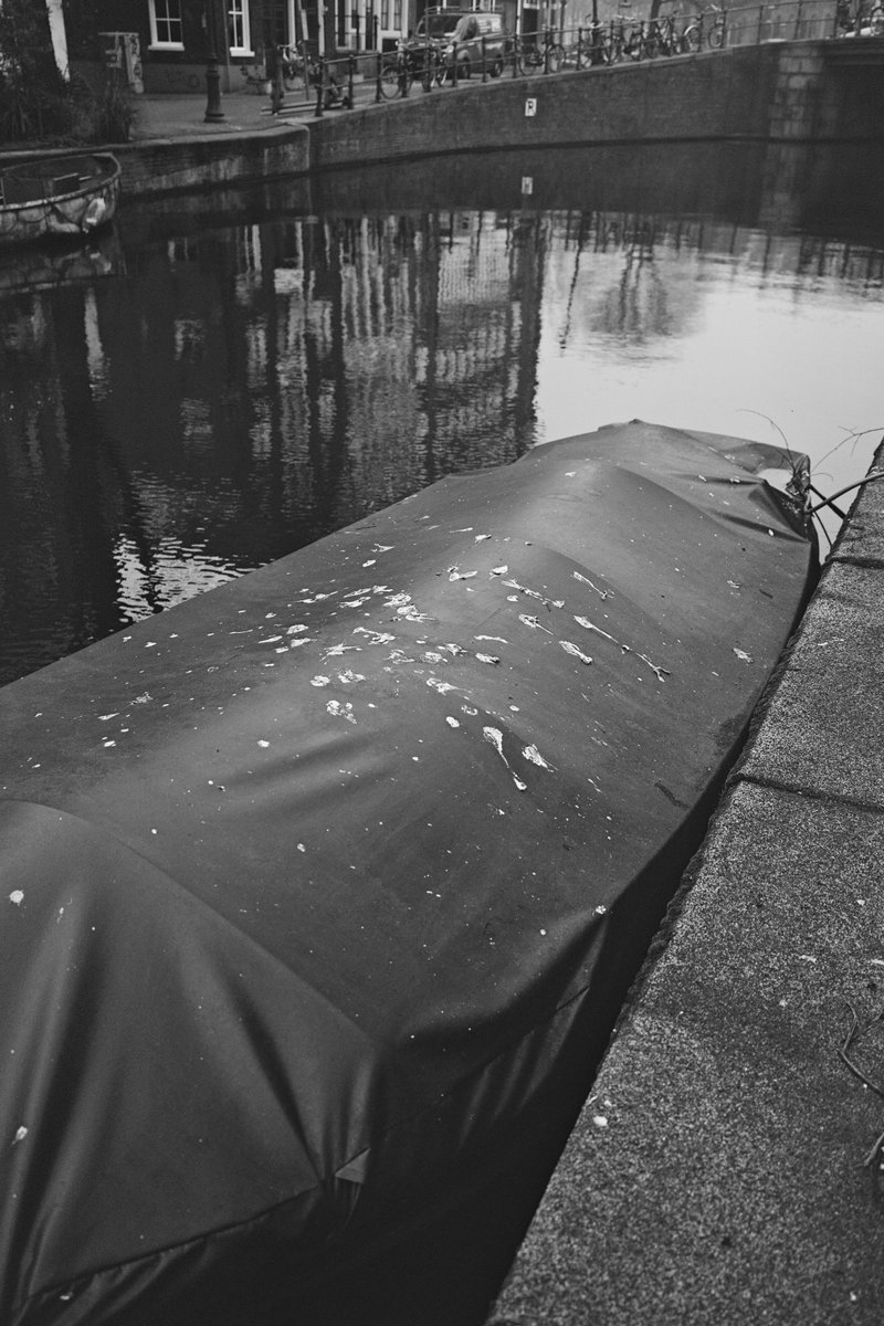 The image shows a small boat moored on a body of water at night, with reflections from lights creating patterns on the water's surface. The boat is partially covered by a black tarp. The setting appears calm and serene, with no people or other significant objects visible in the immediate surroundings.