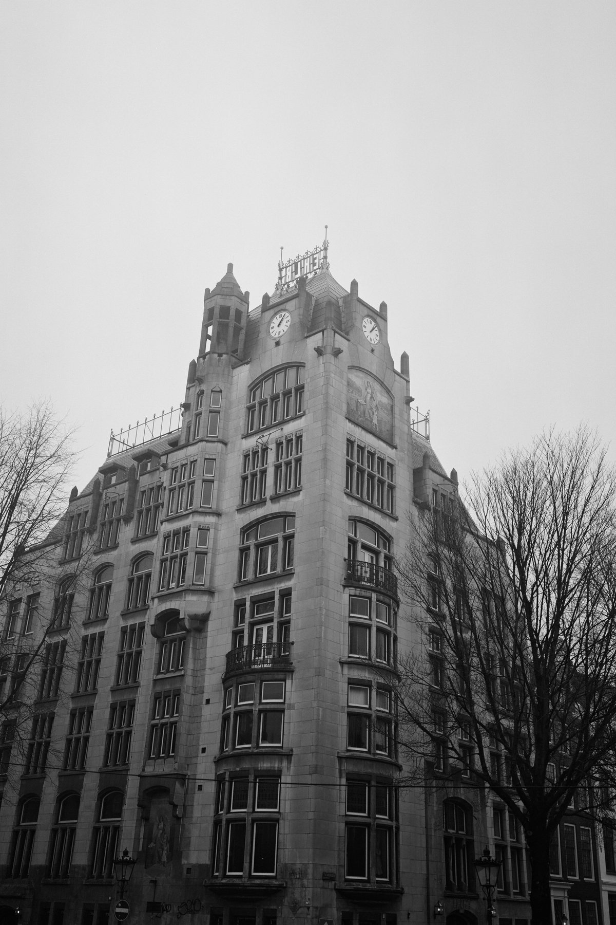  The image shows a tall building with multiple windows and a clock tower. The structure appears modern with a distinctive pattern of glass panels. In the foreground, there are bare trees, suggesting it might be winter or early spring. The photo has an artistic quality, possibly due to filters applied during editing. 