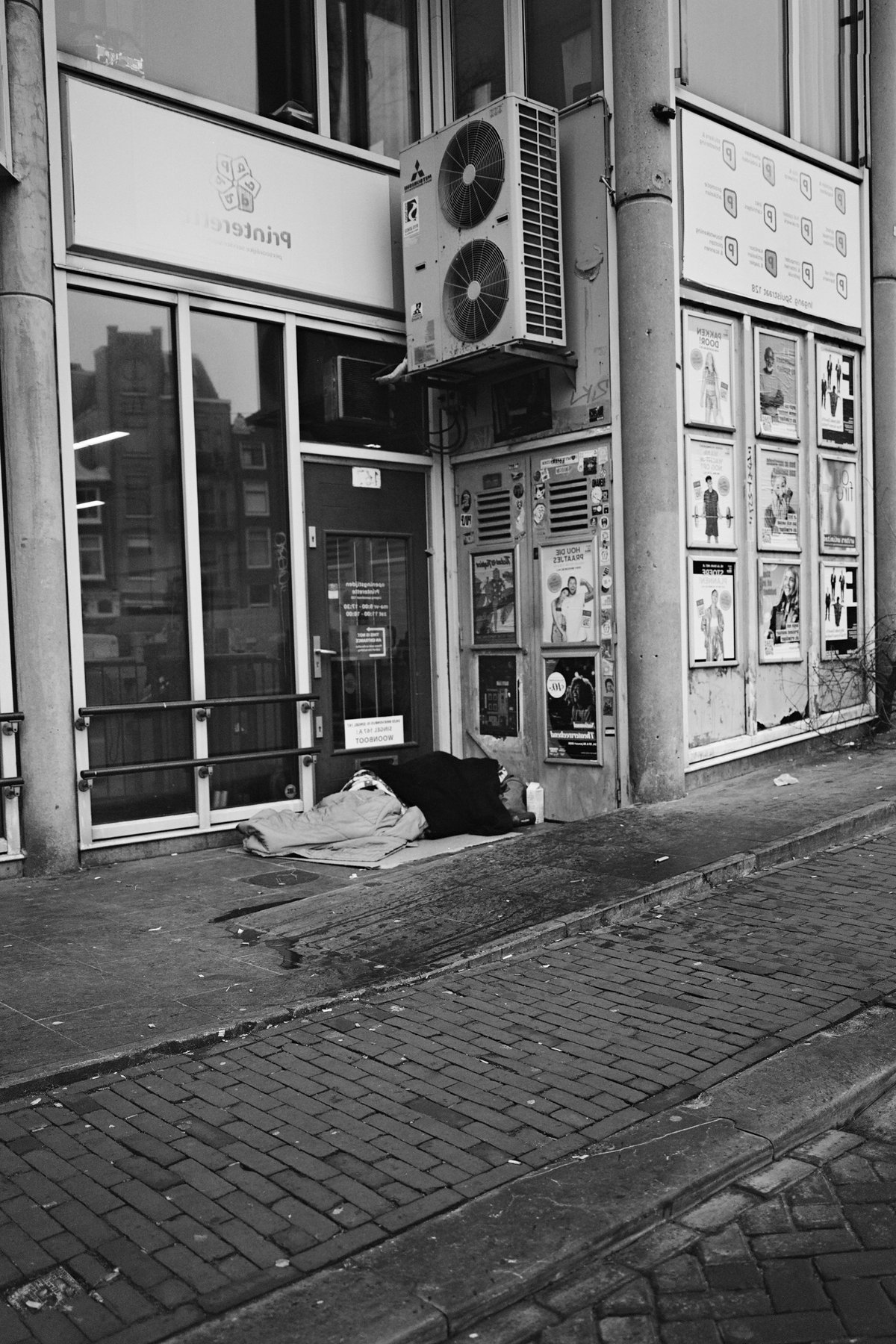  The image shows an urban scene featuring a small business establishment. At the forefront, there's a building with a prominent sign, possibly indicating the name of the shop. An air conditioning unit is visible above the storefront, and there are shelves or display cases inside the store that suggest retail activity. To the left of the image, partially visible, appears to be a person sitting on what seems to be an outdoor step. The street next to the building is empty, with no visible traffic or pedestrians. There's a solitary figure at the far end of the sidewalk, but it's too distant and indistinct to provide details. The overall atmosphere appears calm and quiet, with no evident signs of movement or activity. 